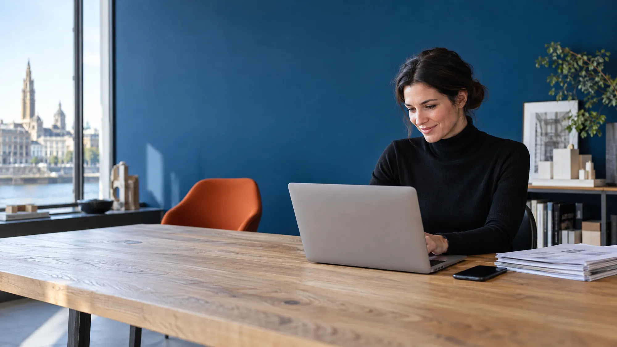 Ondernemer aan haar tafel in een architectenbureau, met laptop en facturen, tegen een kobaltblauwe muur